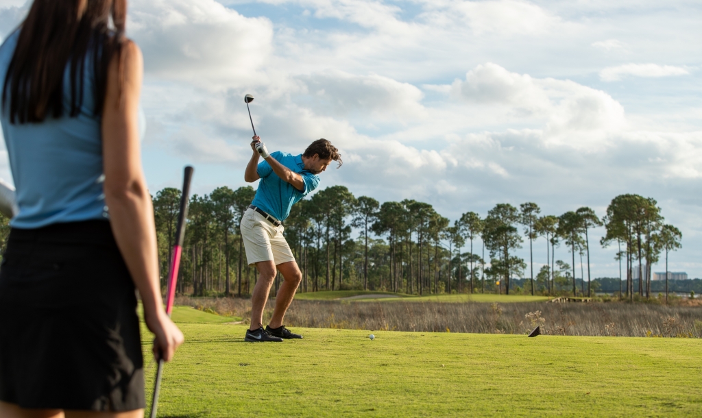 woman holding a golf club with a man teeing off in the background